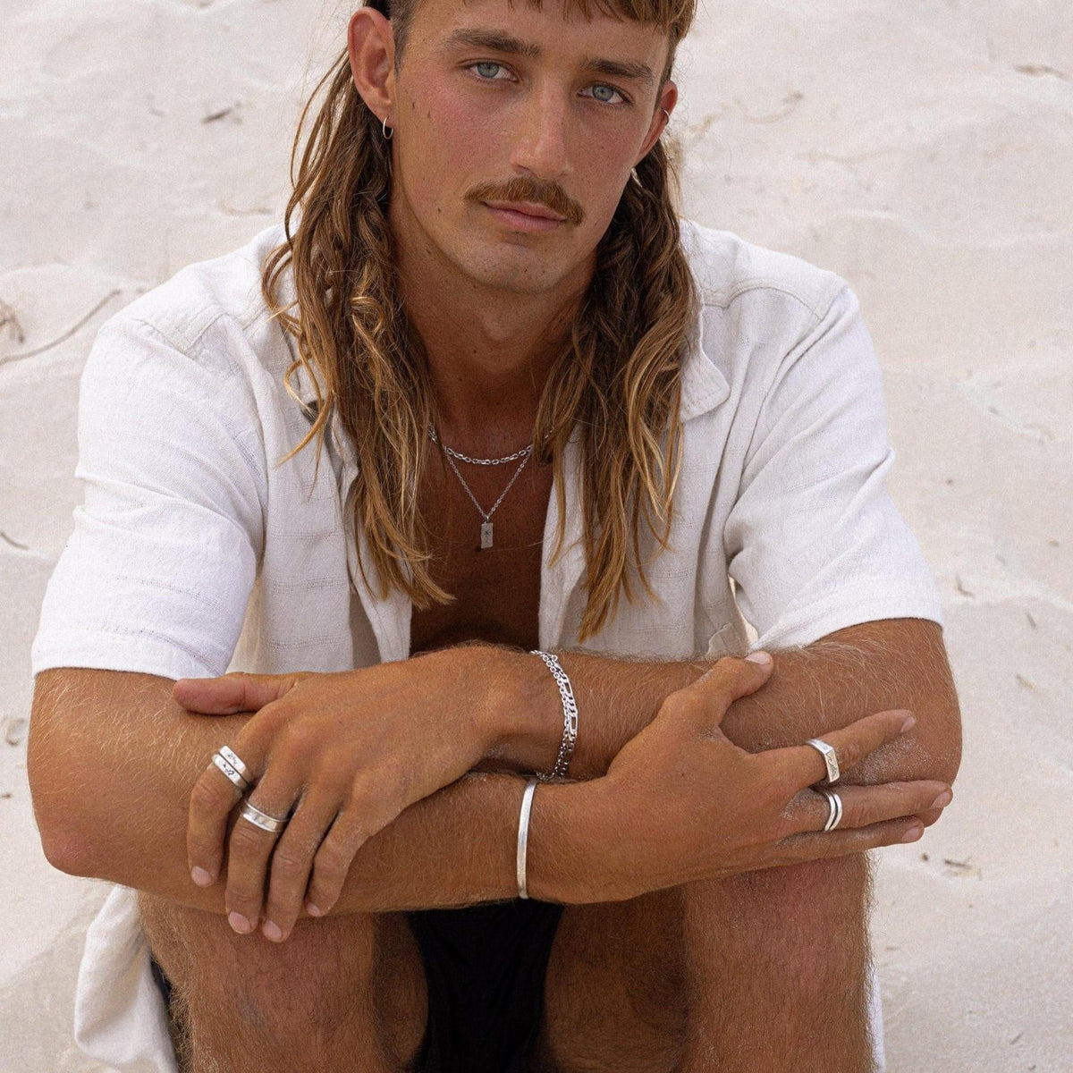 A man models the Billie Jo Drifter & Onyx silver set on a sandy beach, wearing the sterling silver cuff and black onyx ring. A modern, masculine look.