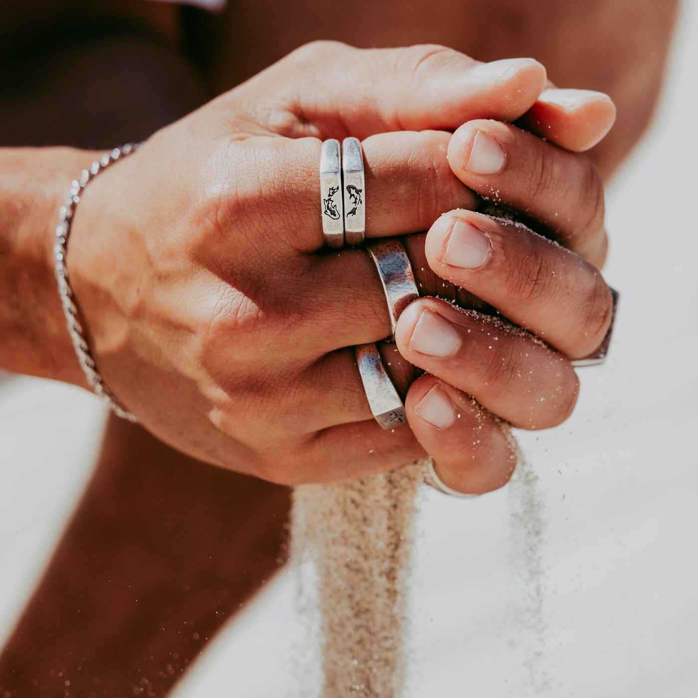 A man's hands covered in sand, wearing the sterling silver Billie Jo KOI I Ring alongside other silver rings, in a warm, sunlit desert setting.