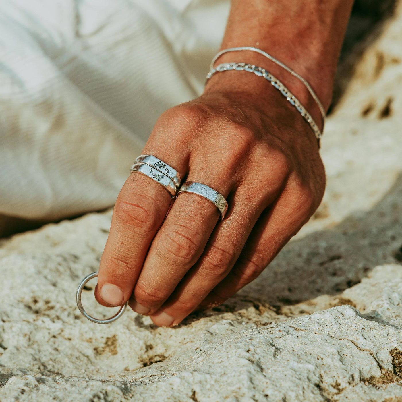 A sun-kissed editorial shot featuring a man's hand adorned with the Billie Jo Serenity Silver ring and other silver jewellery, resting on a coastal rock.
