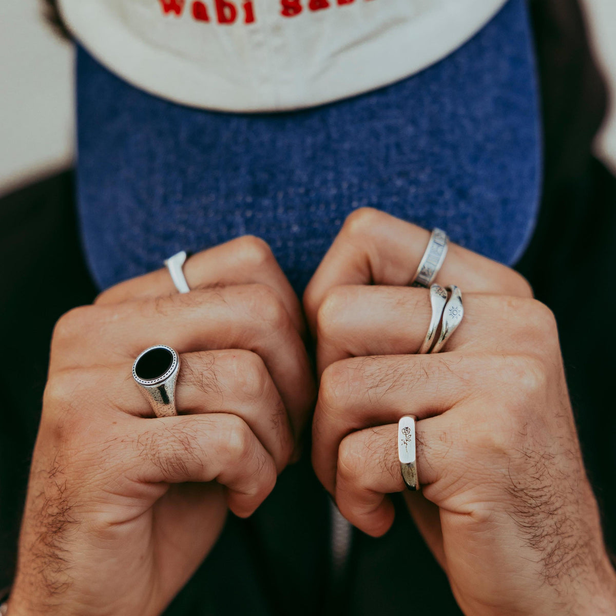 A man wearing a cap with his fists forward, showing off a selection of Billie Jo rings. The Rosa II ring is on his right hand, part of a cool, casual jewellery look.