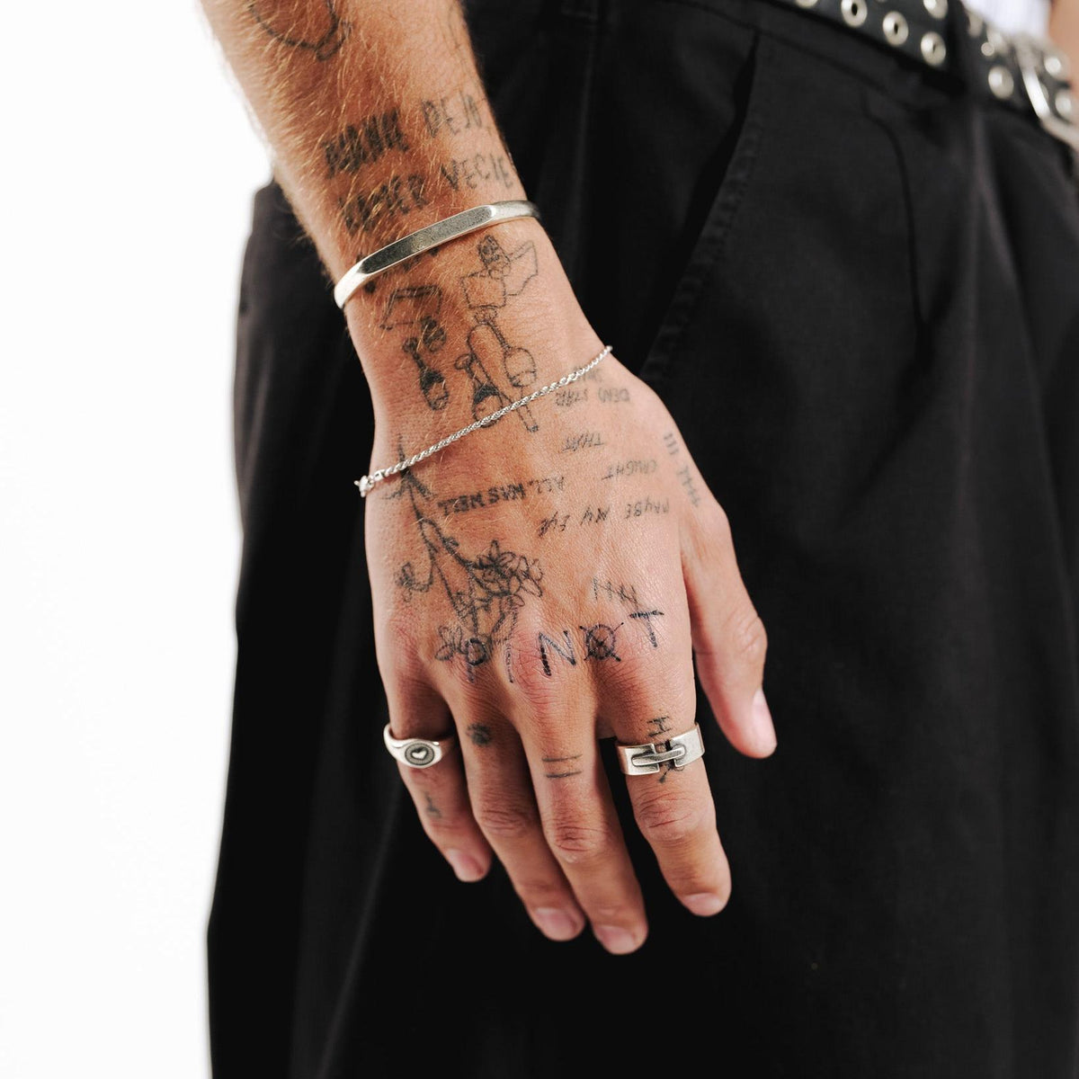 A tattooed man's arm wearing the sterling silver Slim Rope Bracelet from Billie Jo, styled with a silver cuff and rings against black trousers.