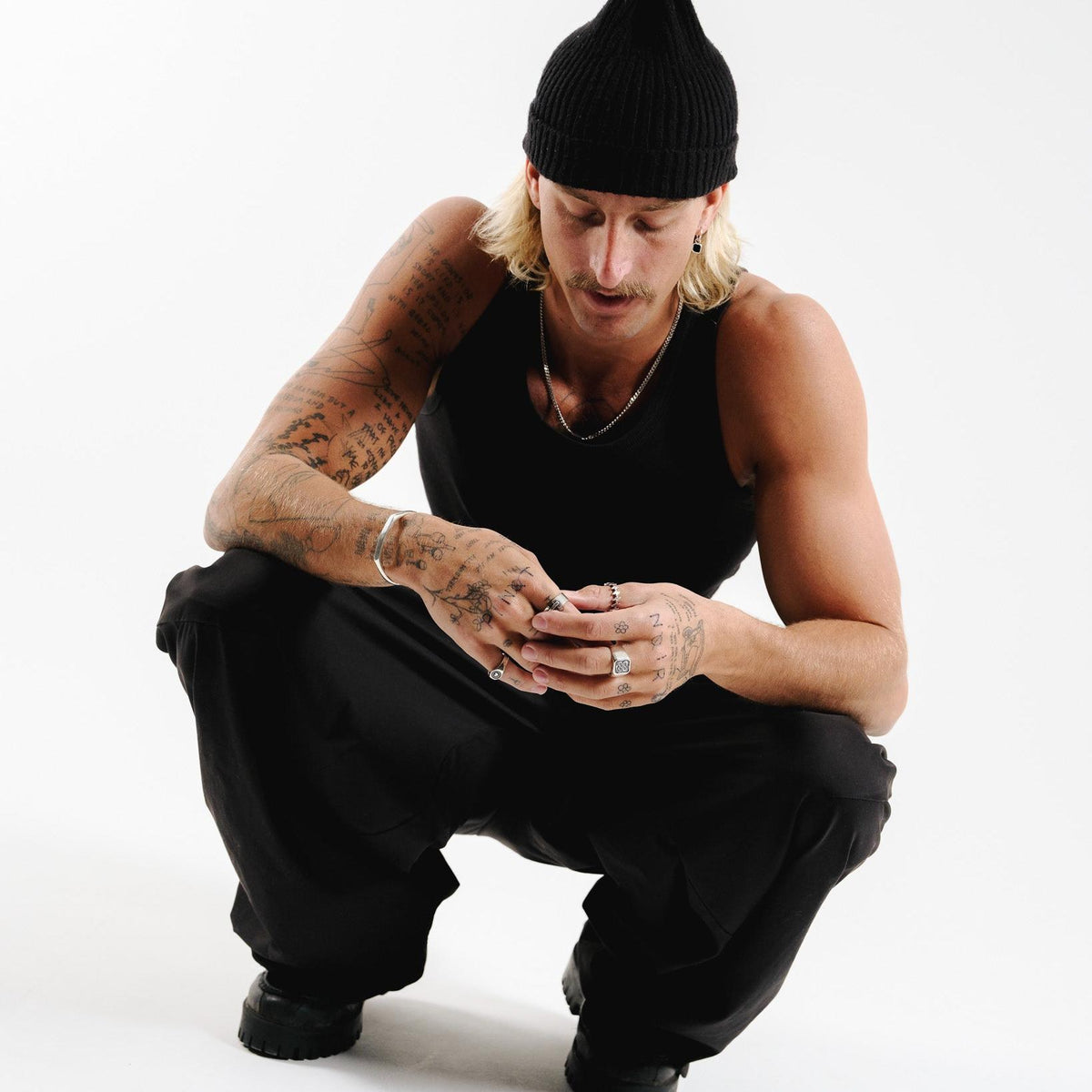 A model with blonde hair and a black beanie crouches, his tattooed hands clasped, wearing multiple silver rings including the Vita Ring by Billie Jo. This editorial shot captures the brand's modern aesthetic.