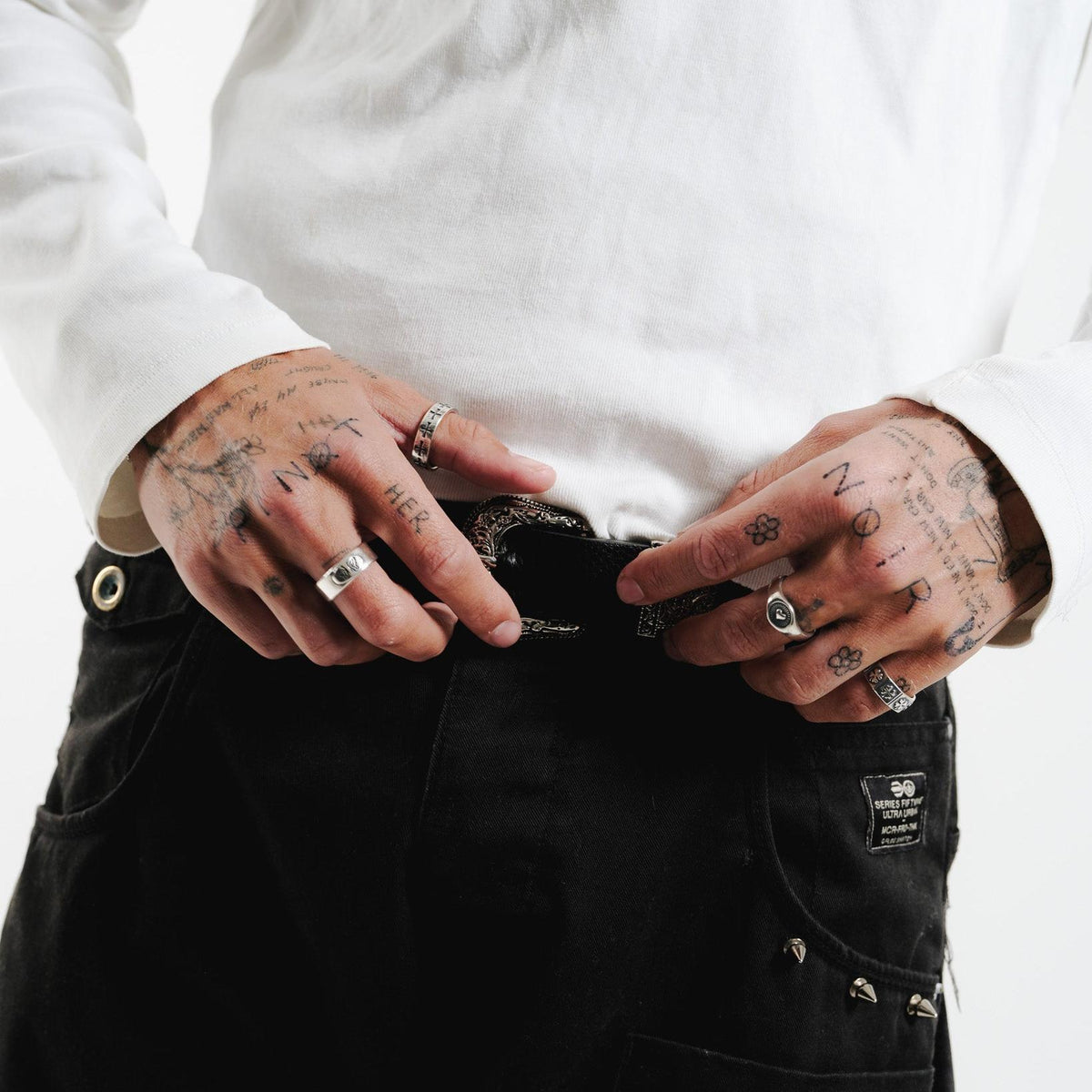 A close-up of a man's hands with tattoos, wearing the Billie Jo Sanctum Ring and other silver jewellery. He is adjusting a decorative belt buckle.