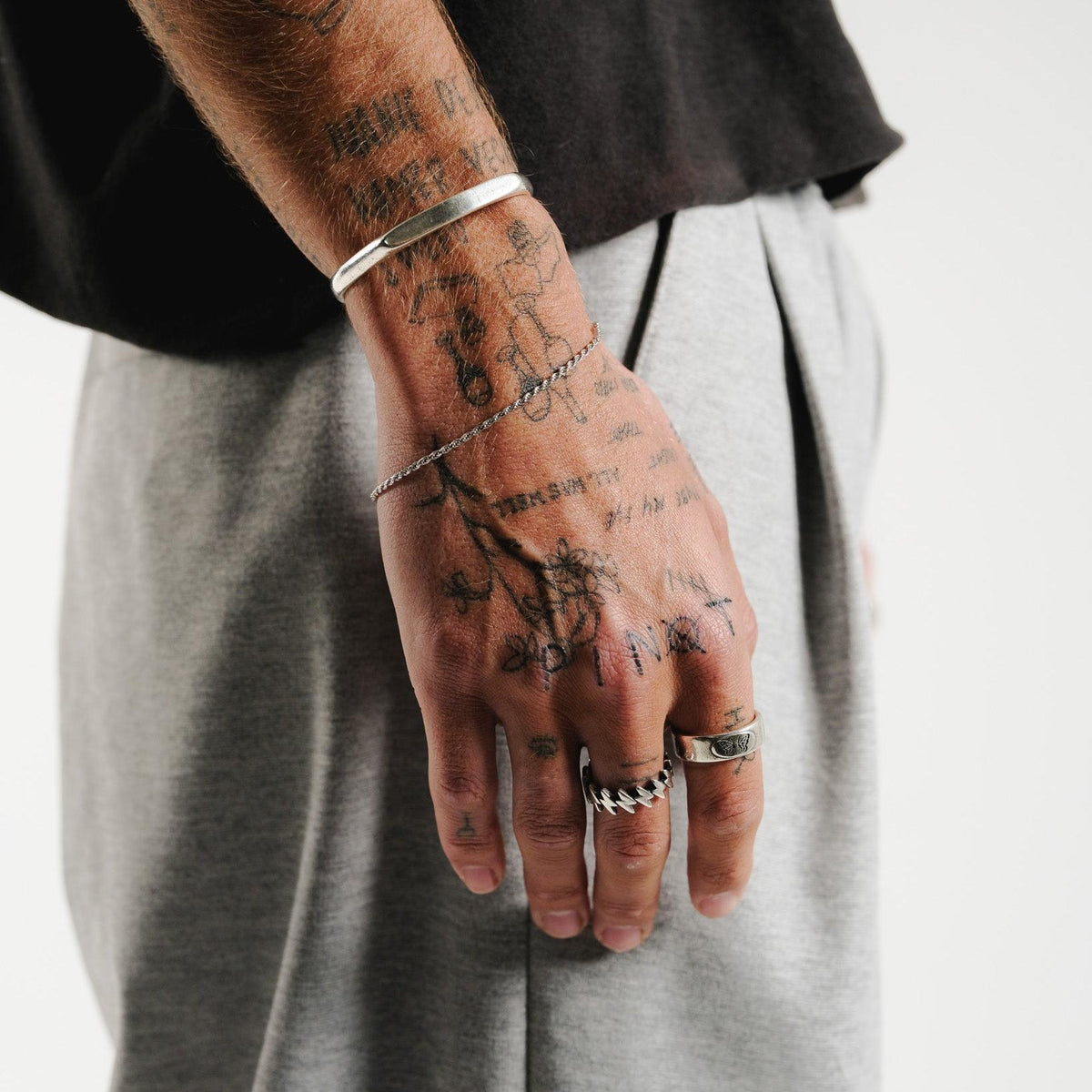 On a tattooed arm, the sterling silver Echo Cuff by Billie Jo is styled with a chain bracelet and rings. A casual look with a black tee and grey trousers, showcasing versatile men's jewellery.
