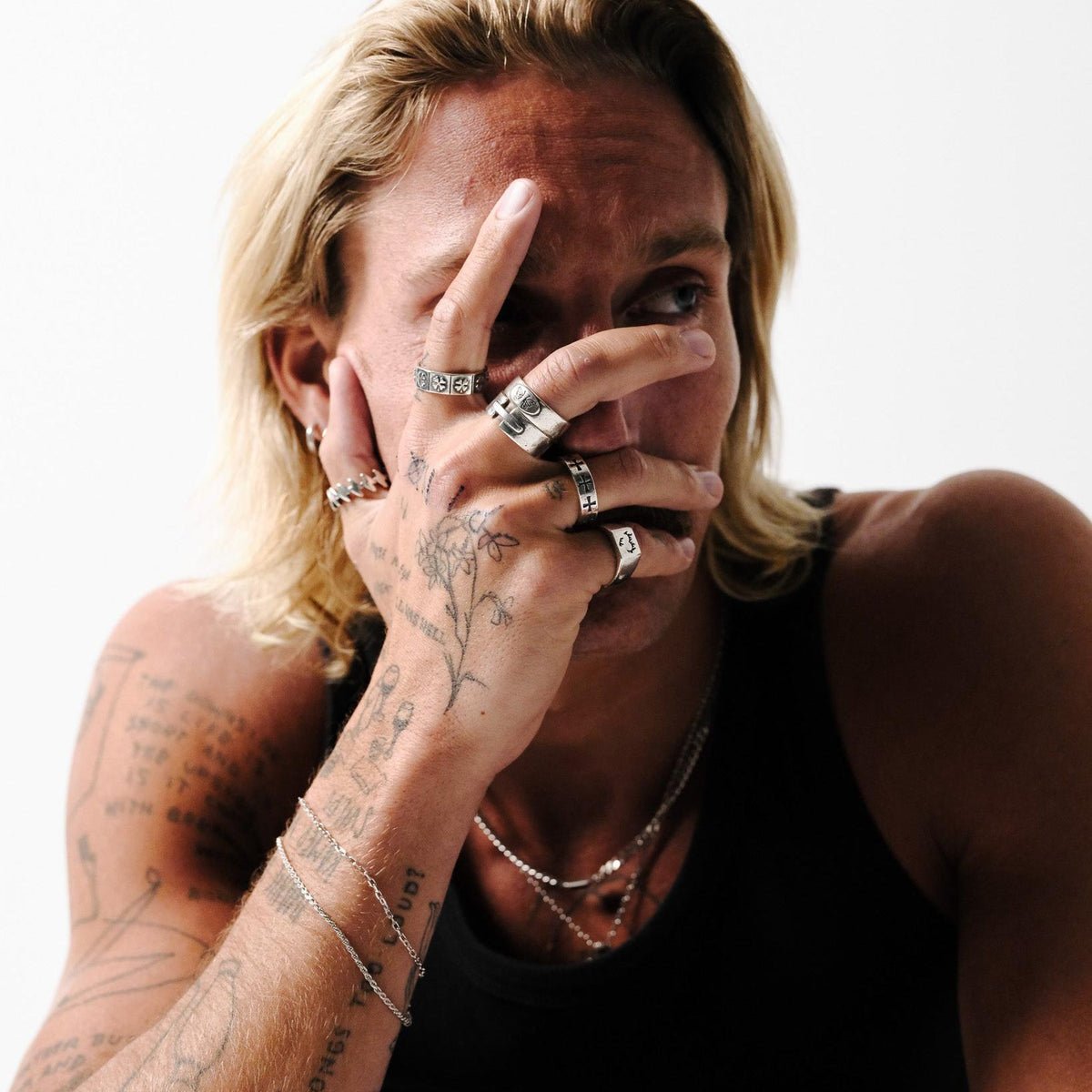 A male model with long blonde hair and tattooed arms, wearing the Billie Jo Slim Batch Bracelet with a stack of silver rings and necklaces for a modern editorial look.