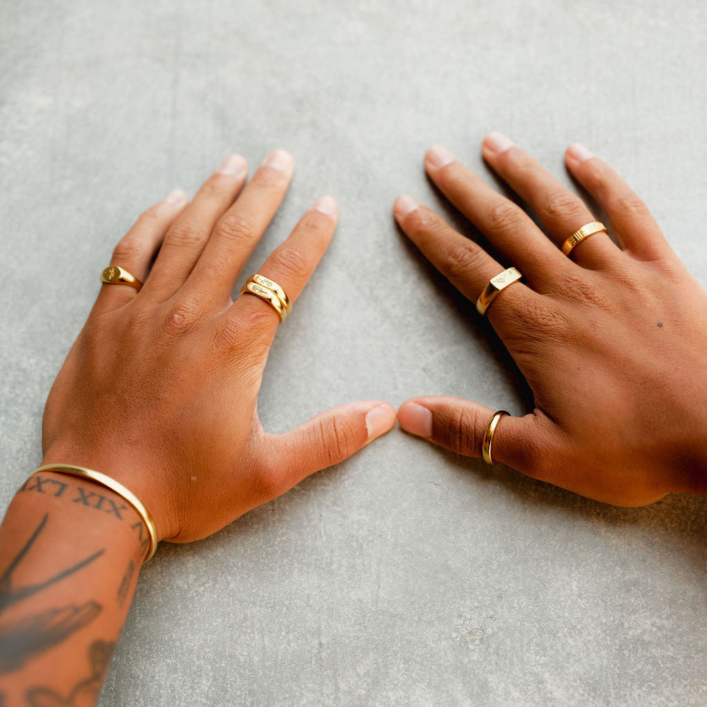 A top-down view of a man's hands with tattoos, showcasing a curated selection of Billie Jo's gold rings, including the Stacker Ring, on a grey concrete-style background.