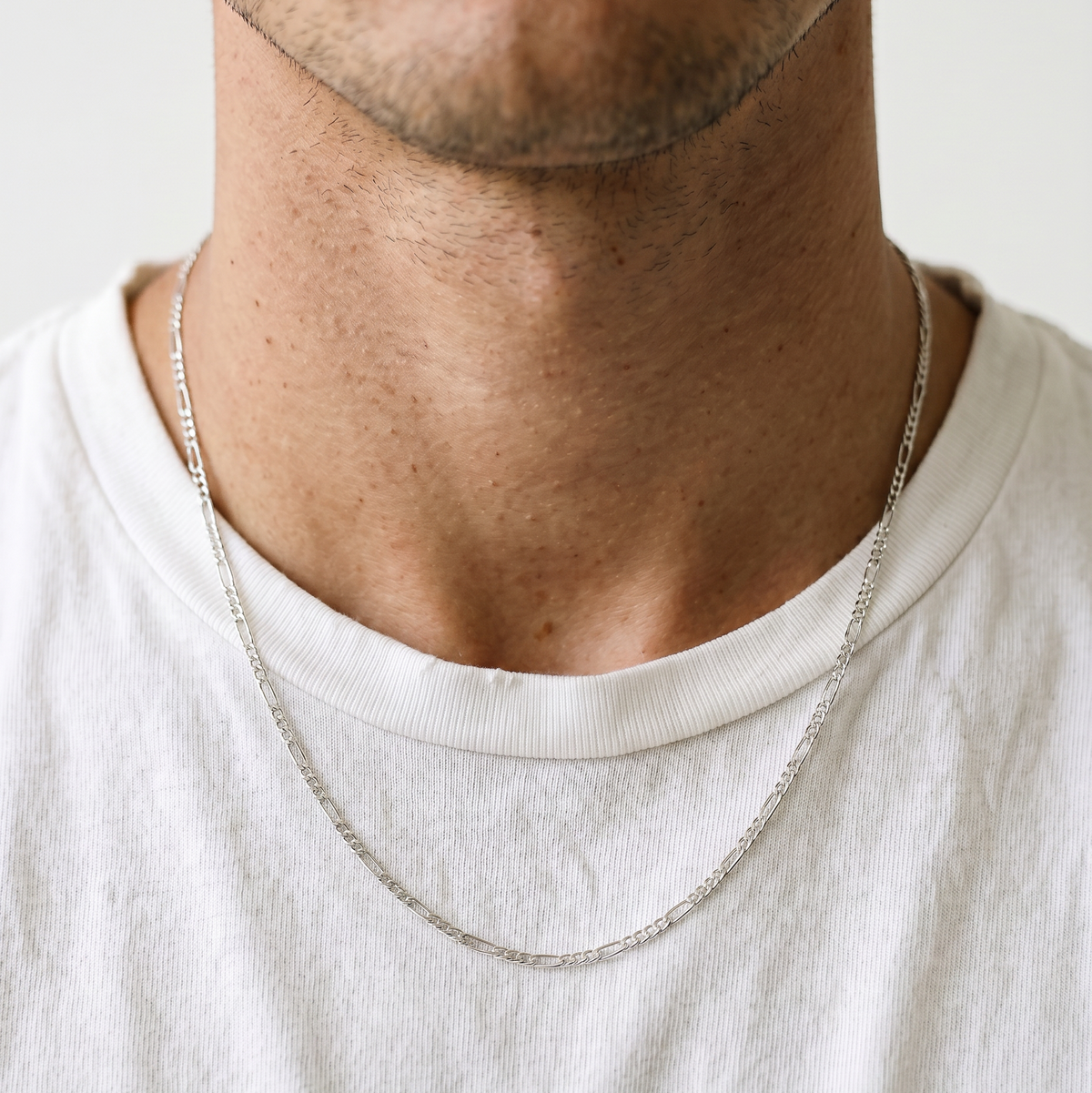 A male model wearing a gold tone stainless steel slim Figaro chain necklace from Australian jewellery brand Billie Jo, shot against a sleek silver chrome backdrop showcasing the delicate alternating link design perfect for everyday wear.