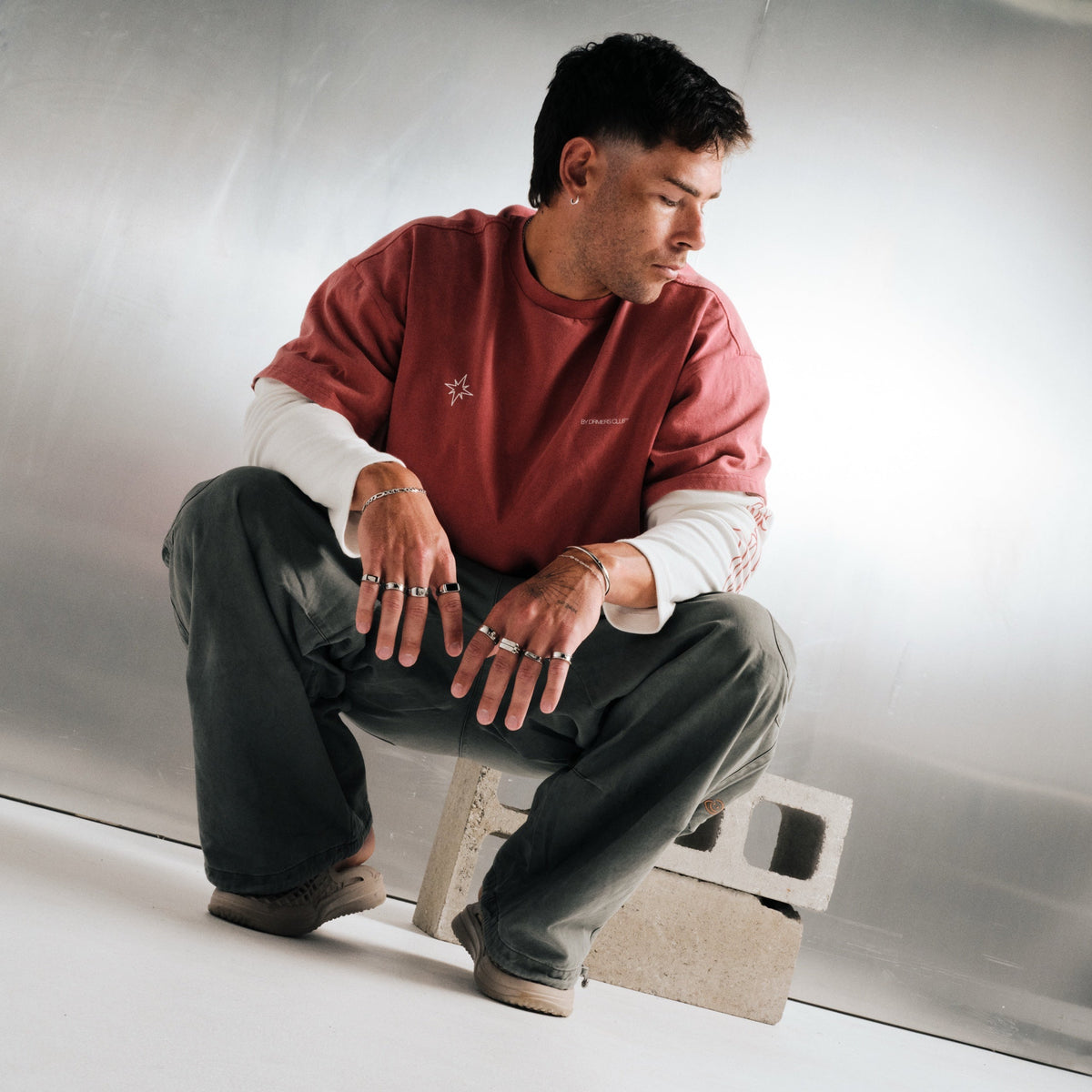 Man wearing silver finish stainless rings, crouched on a concrete block, with slim steel bracelets
