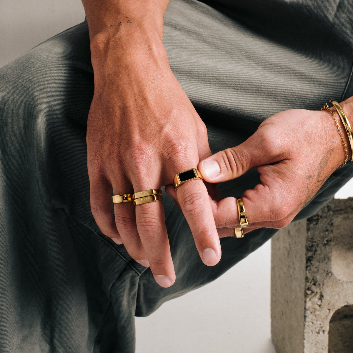 Men's Gold Forge Ring in 18k gold infused stainless steel, worn on the ring finger, seated, hands together over his knee