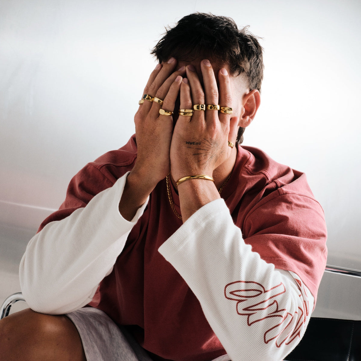 Man wearing gold stainless rings, hands over his face, with a gold chain necklace, gold bangle