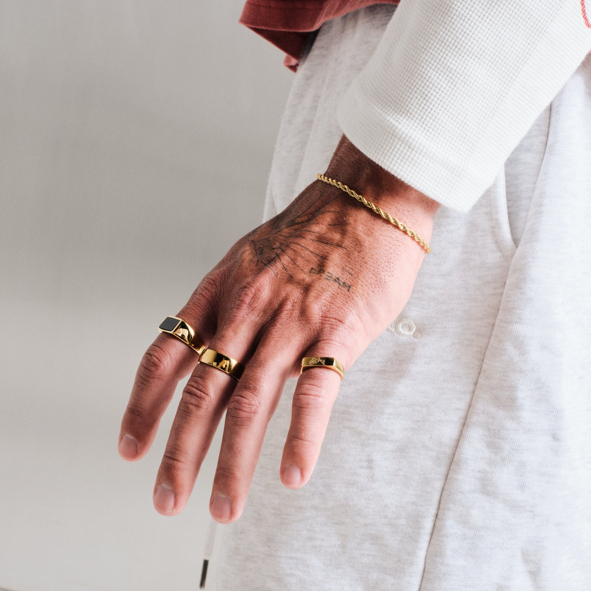 Men's Gold Koi II Ring in 18k gold infused stainless steel, in a stack, hand hanging by his side, with a gold rope bracelet