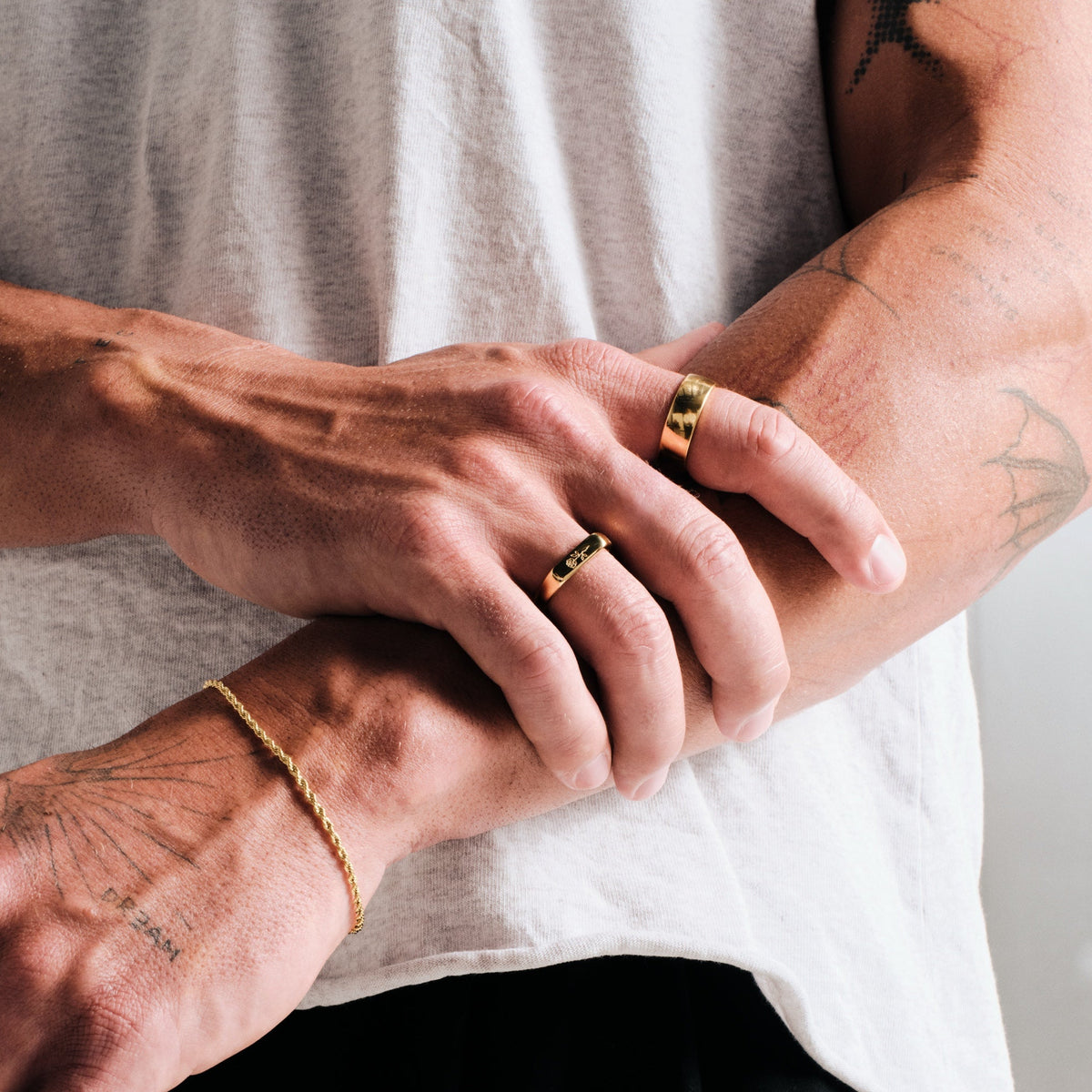 Men's Gold Rosa II Ring in 18k gold infused stainless steel, in a stack, arms folded, with a gold rope bracelet on left wrist