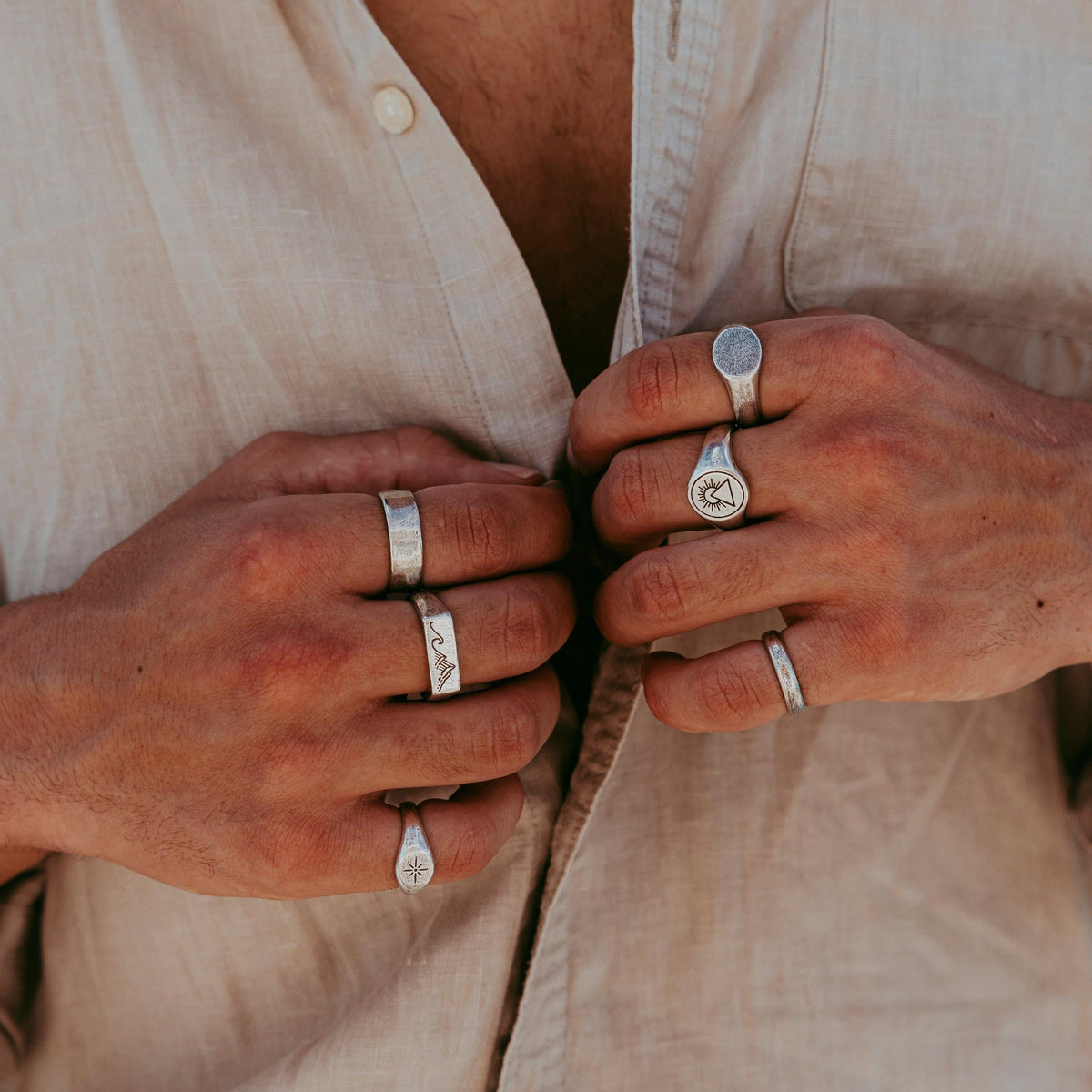 A man wearing multiple sterling silver rings from Billie Jo, including the Luminous Ring on his pinky finger, while buttoning a cream linen shirt.