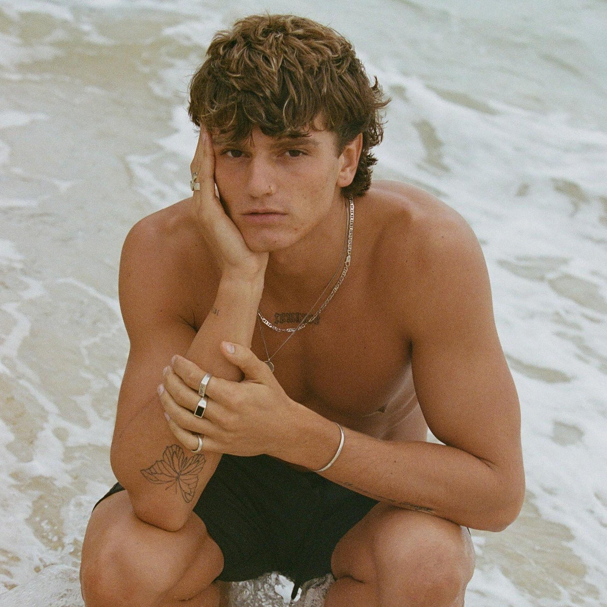 A young man with curly hair sits in the ocean surf wearing the Billie Jo Mariner Necklace, embodying a cool and carefree Australian vibe.