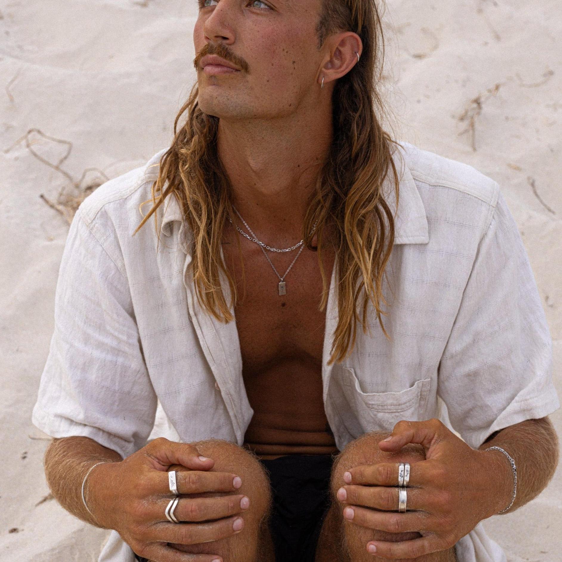 A man with long hair and a mustache sits on sandy ground, wearing a light-colored, short-sleeved shirt, multiple rings, and layered necklaces, looking slightly upward with a thoughtful expression.