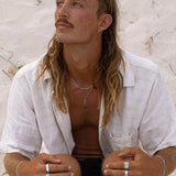 A man with long hair and a mustache sits on sandy ground, wearing a light-colored, short-sleeved shirt, multiple rings, and layered necklaces, looking slightly upward with a thoughtful expression. v2