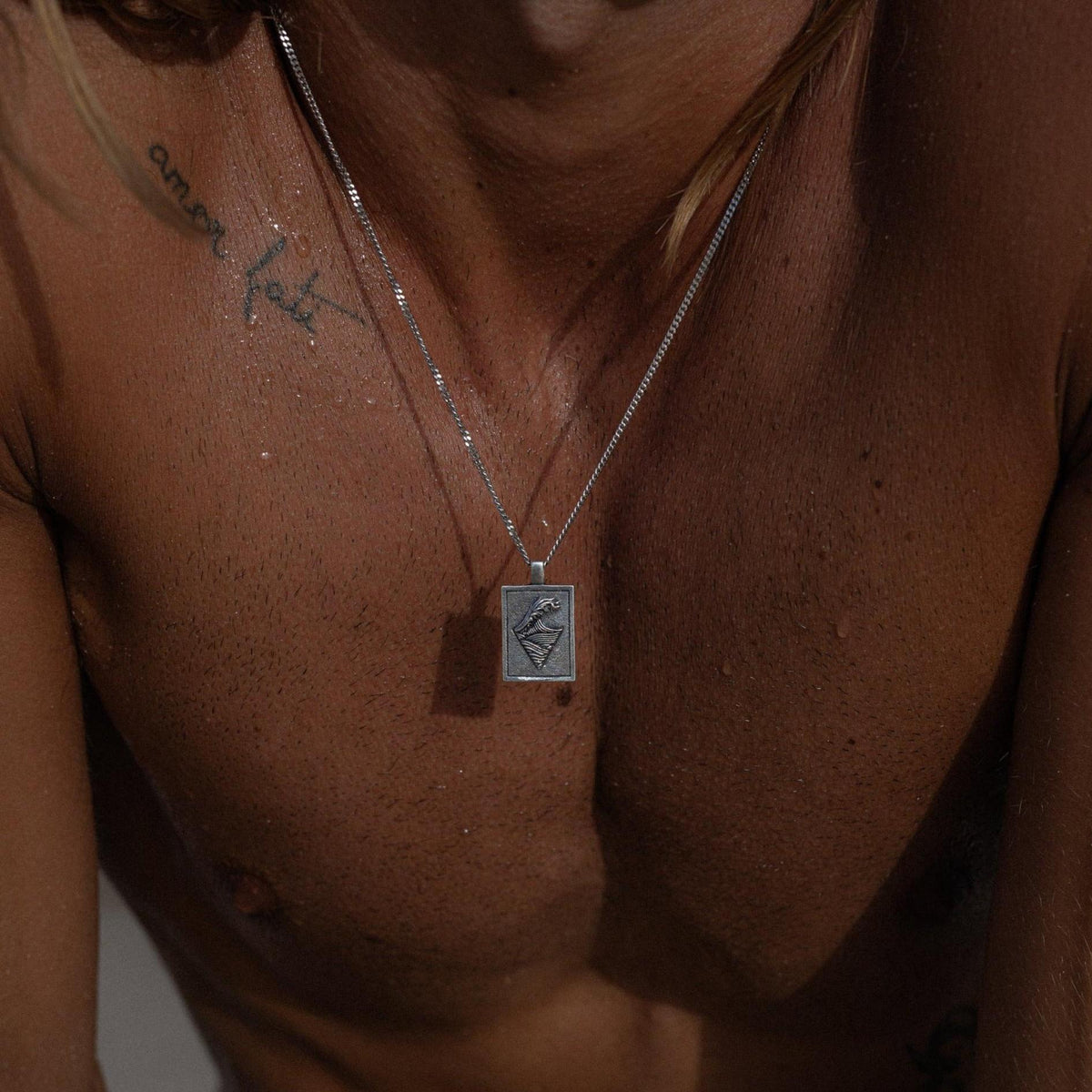 A man wearing the sterling silver Burleigh Pendant by Billie Jo, the rectangular pendant with its wave engraving resting on his chest in a moody, editorial shot.