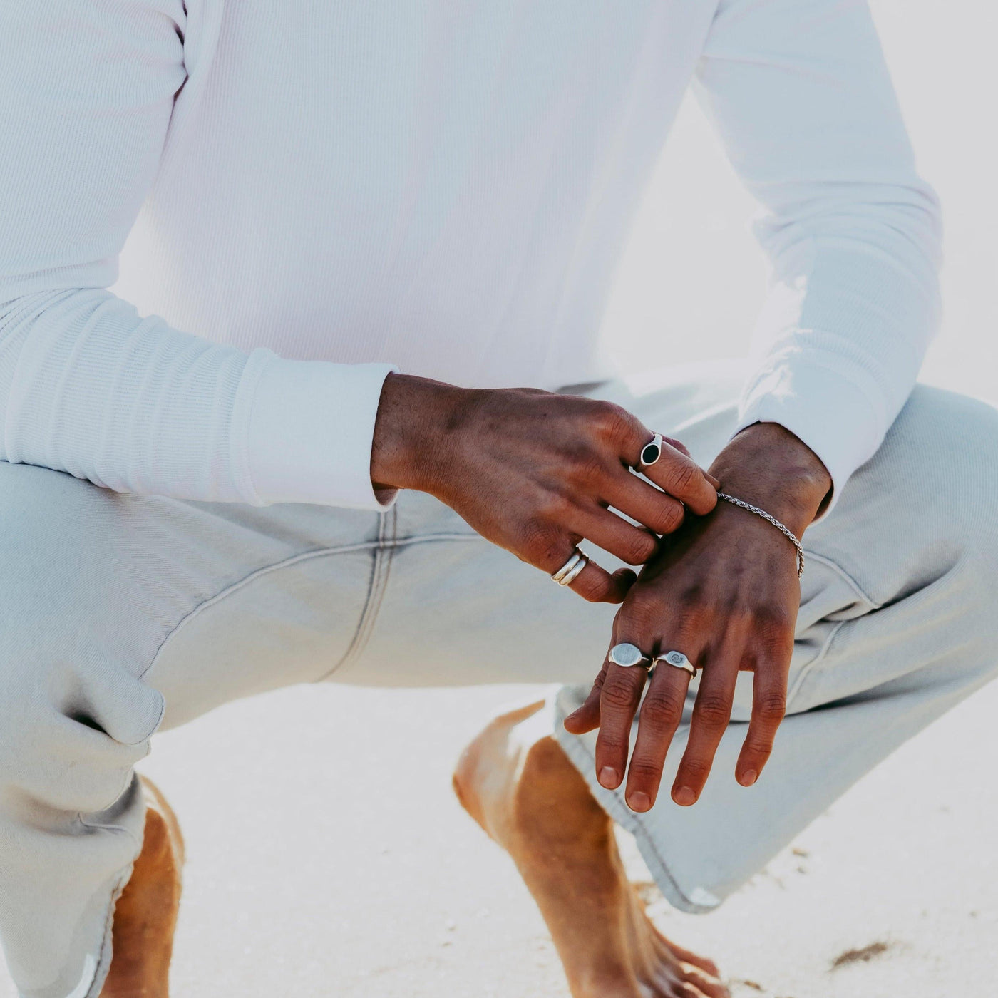 A man crouching on a sandy beach, wearing a white long-sleeved top and light-coloured jeans, with the Billie Jo Signet Ring on his hand.