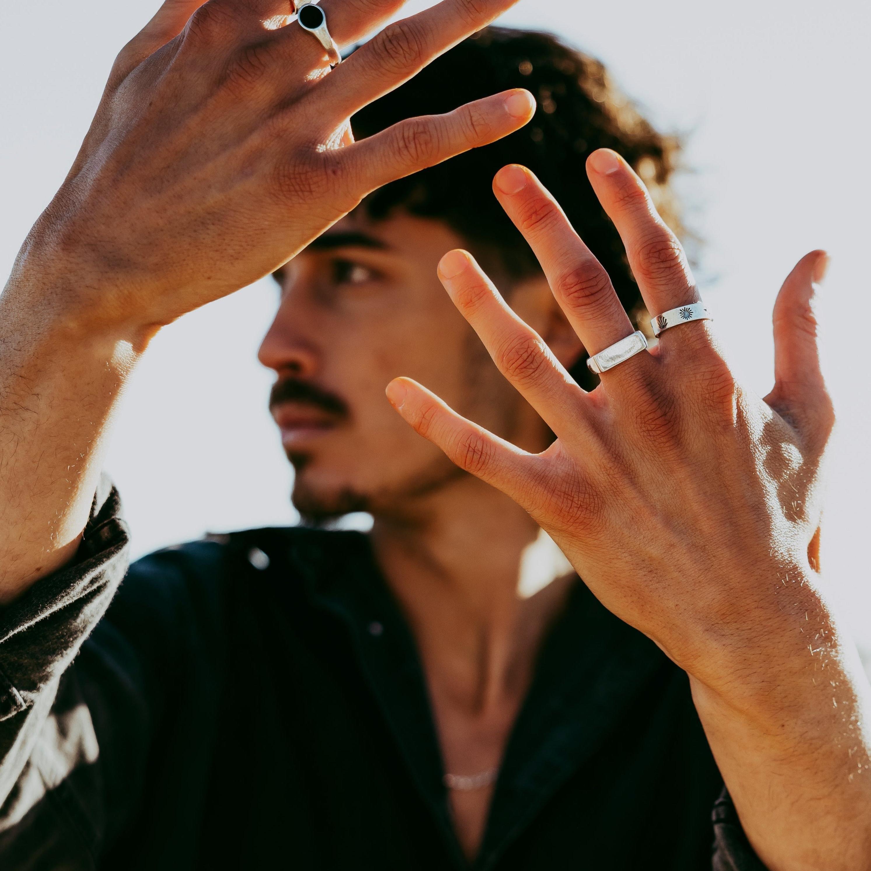 A man with dark curly hair and a moustache holds his hands up to his face, shielding from the sun. He wears the sterling silver Elation Ring from Billie Jo.