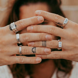 A person holds both hands in front of their face, fingers spread, displaying silver rings and finger tattoos. The background is blurred, highlighting the hands, rings, and tattoos. The person has long hair.