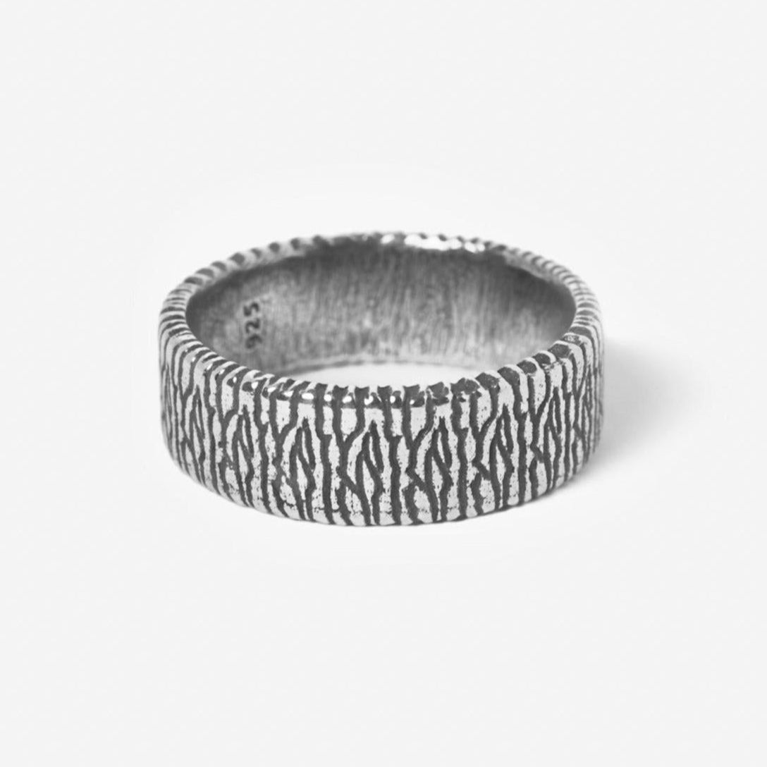 A silver ring with a wide band featuring an engraved, organic, vertical line pattern resembling wood grain, displayed on a plain white background.