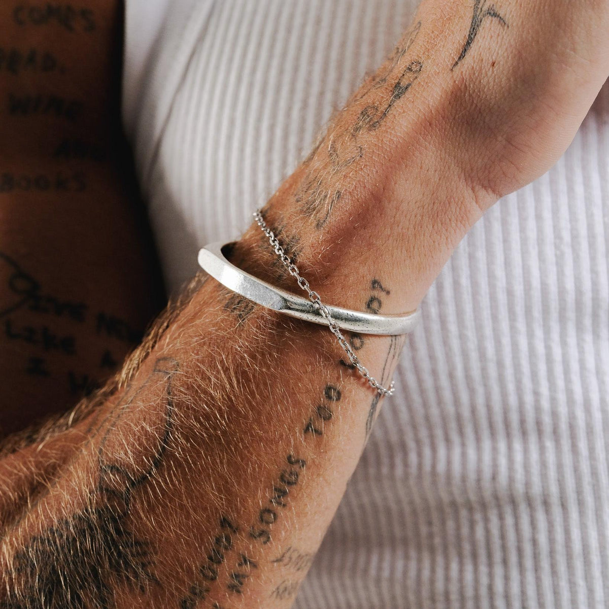 Close-up of the Billie Jo Echo Cuff on a man's tattooed arm, showing the cuff's textured silver finish and how it layers with a delicate chain bracelet. Modern men's jewellery from Australia.