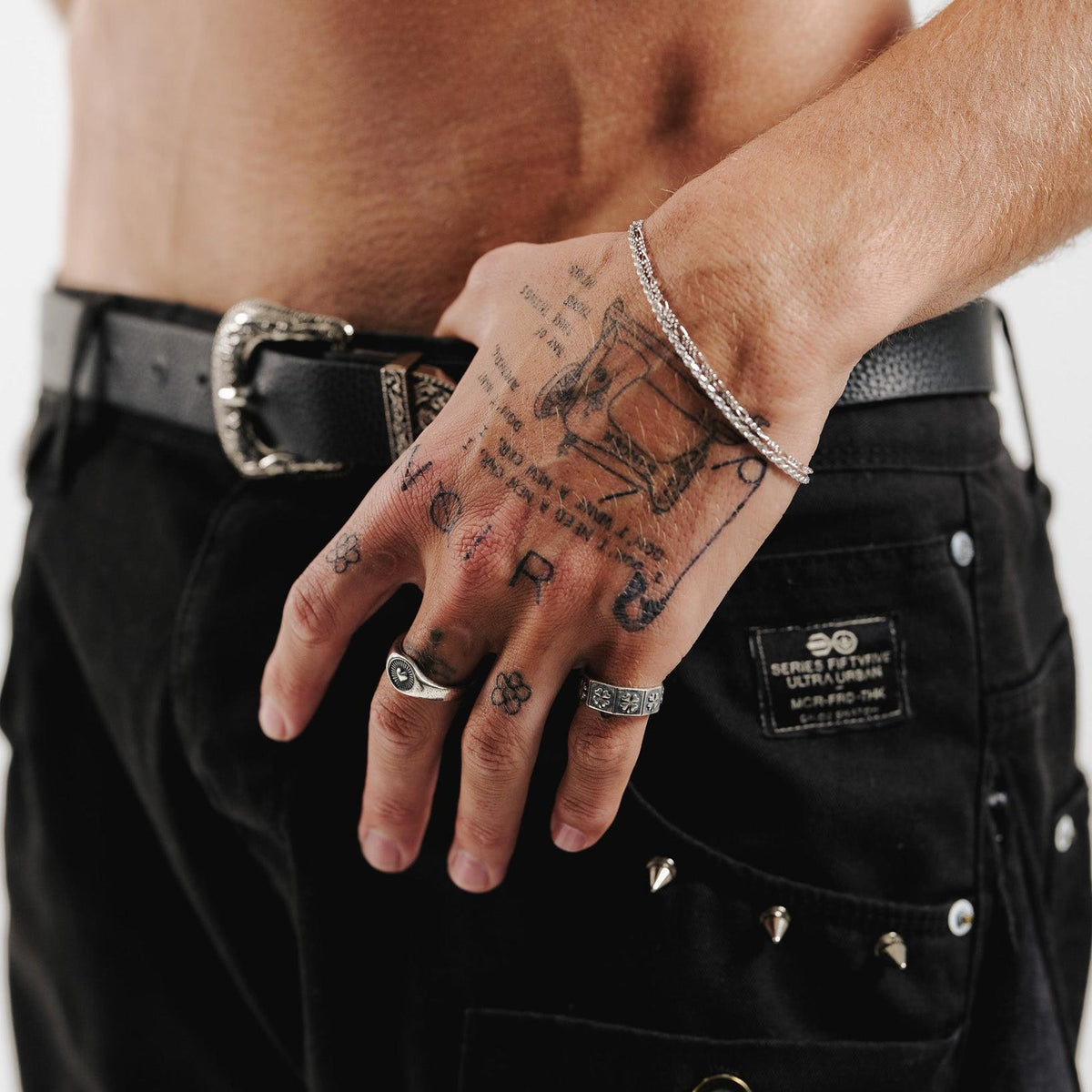 A man with tattooed hands wears the sterling silver Billie Jo LUCKY RING, styled with black jeans and a decorative silver belt buckle for a cool, modern look.