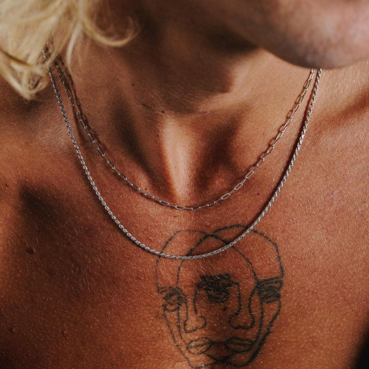 A moody, close-up shot of the Billie Jo Slim Link Necklace in sterling silver, layered on a man's chest with dramatic lighting.
