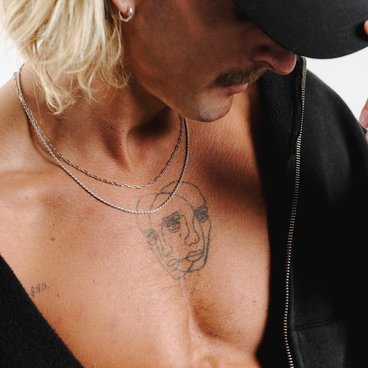 A man with a moustache and blonde hair wears the sterling silver Carve Earring by Billie Jo, styled with layered silver necklaces and a black zip-up hoodie for a modern, edgy look.