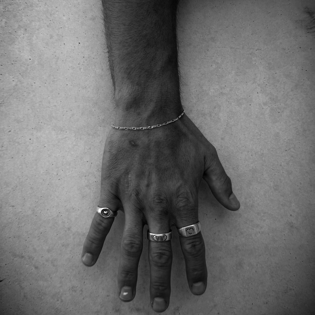 A black and white close-up of a man's hand wearing the Billie Jo Slim Figaro Bracelet and several silver rings. This artistic shot focuses on the jewellery against a textured background, creating a moody and masculine vibe.