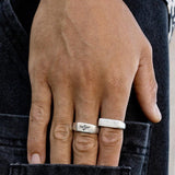 A close-up of a hand wearing two silver rings and a silver bracelet, with fingers resting on the back pocket of dark denim jeans. The hand is partially tucked into the pocket.