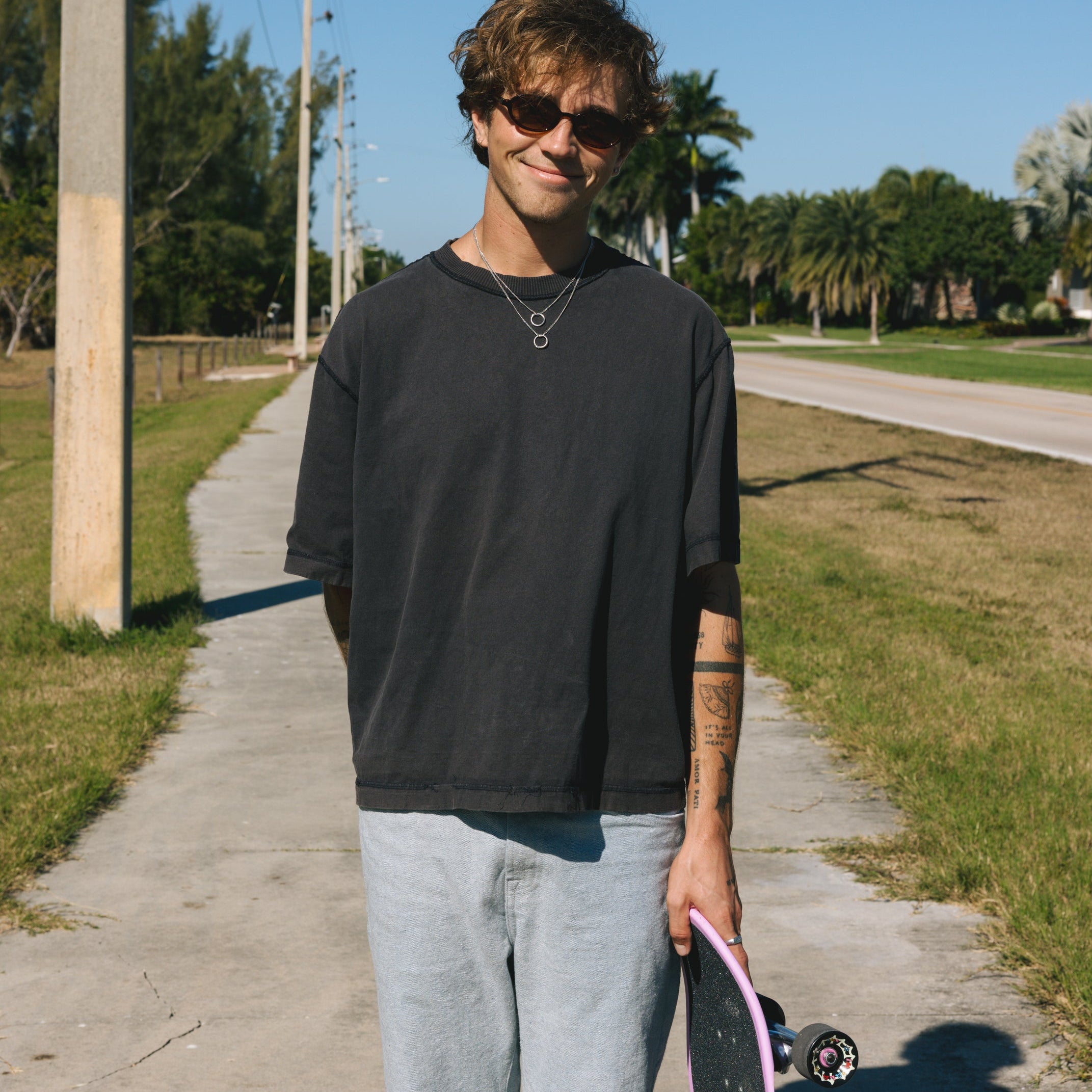 Man holding a skateboard wearing jewellery on a sidewalk with trees in the background