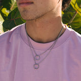 Person wearing a pink shirt with a necklace featuring a snake circular pendant, surrounded by green foliage.