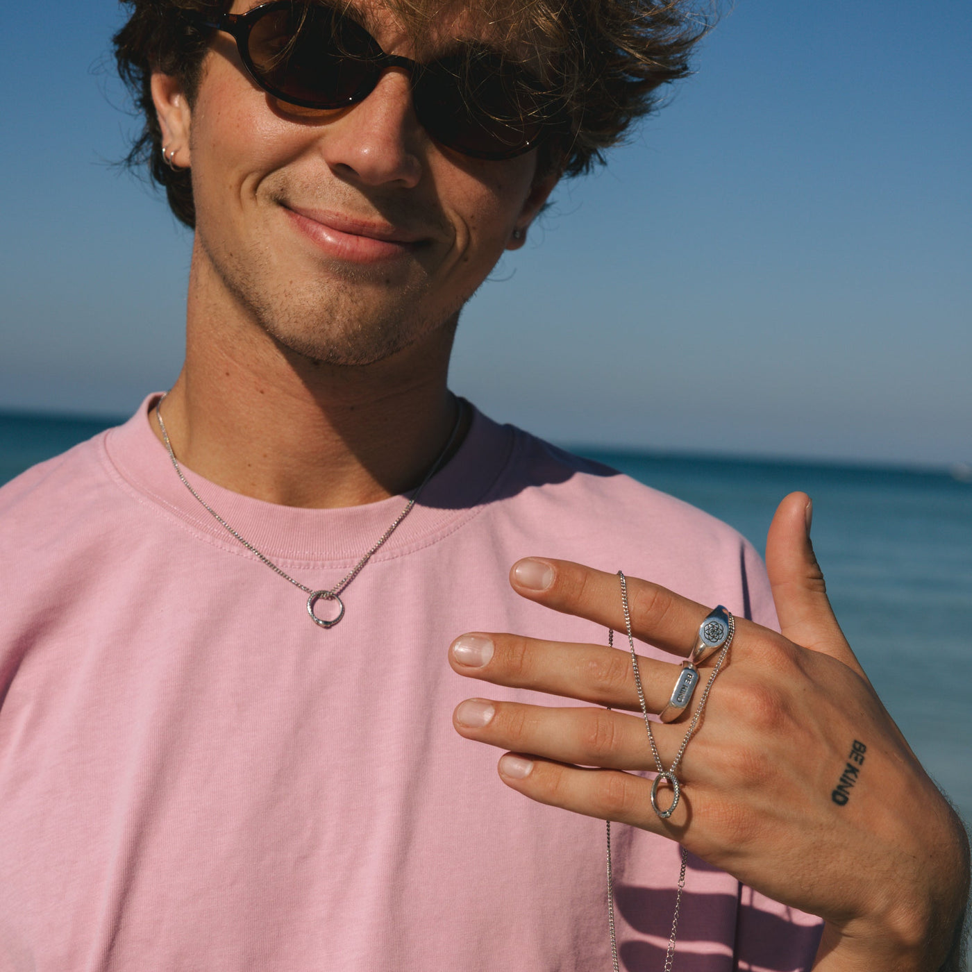A smiling man on a sunny Australian beach wearing a pink t-shirt, sunglasses, and Billie Jo's sterling silver UNITY RING and necklace.