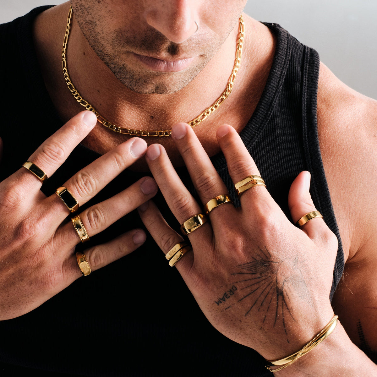 Man wearing gold stainless rings, hands on his chest, with a gold Figaro chain, gold cuff and slim bracelet, black singlet
