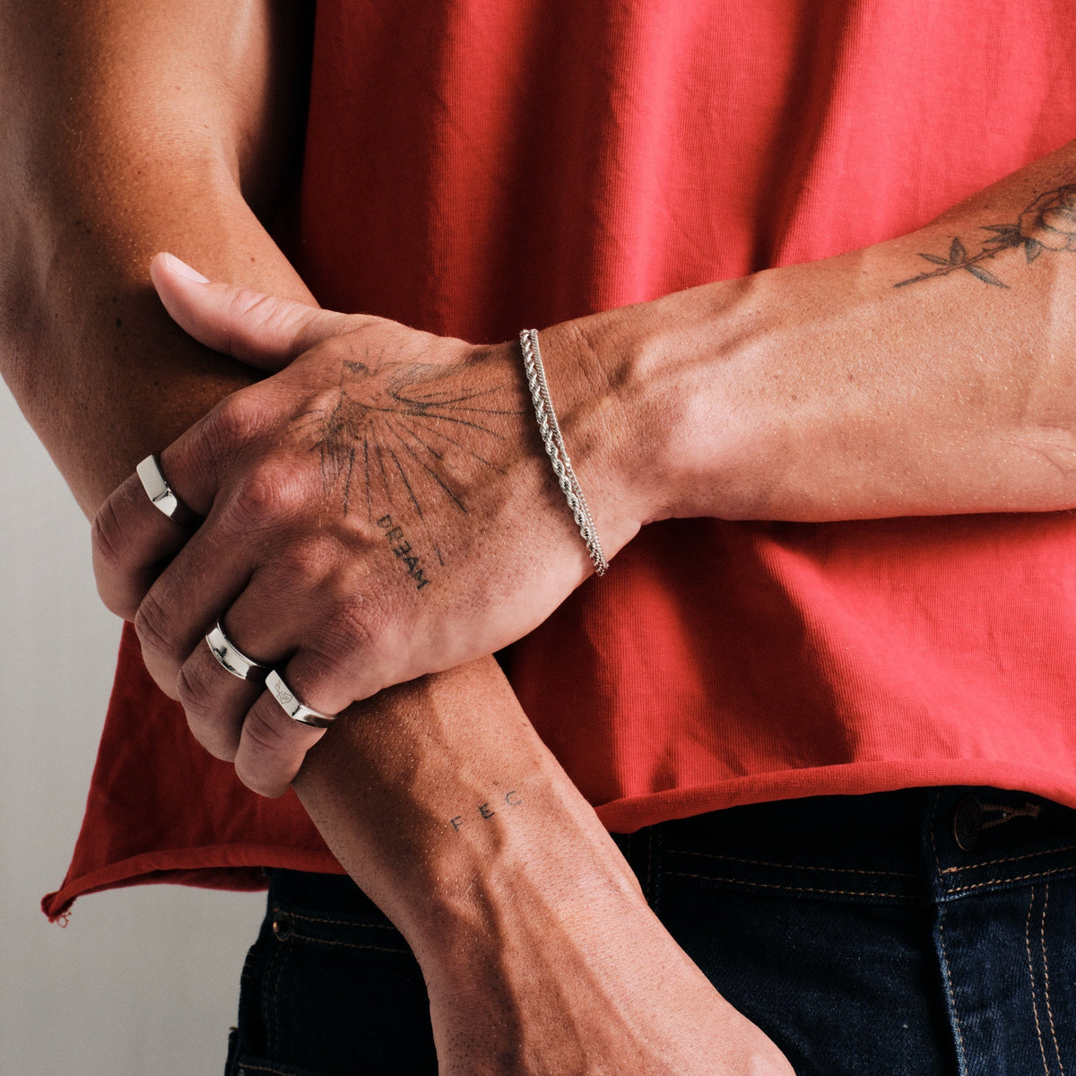 Man wearing silver finish stainless rings, arms crossed at the waist, with a steel rope bracelet, red tee, dark jeans