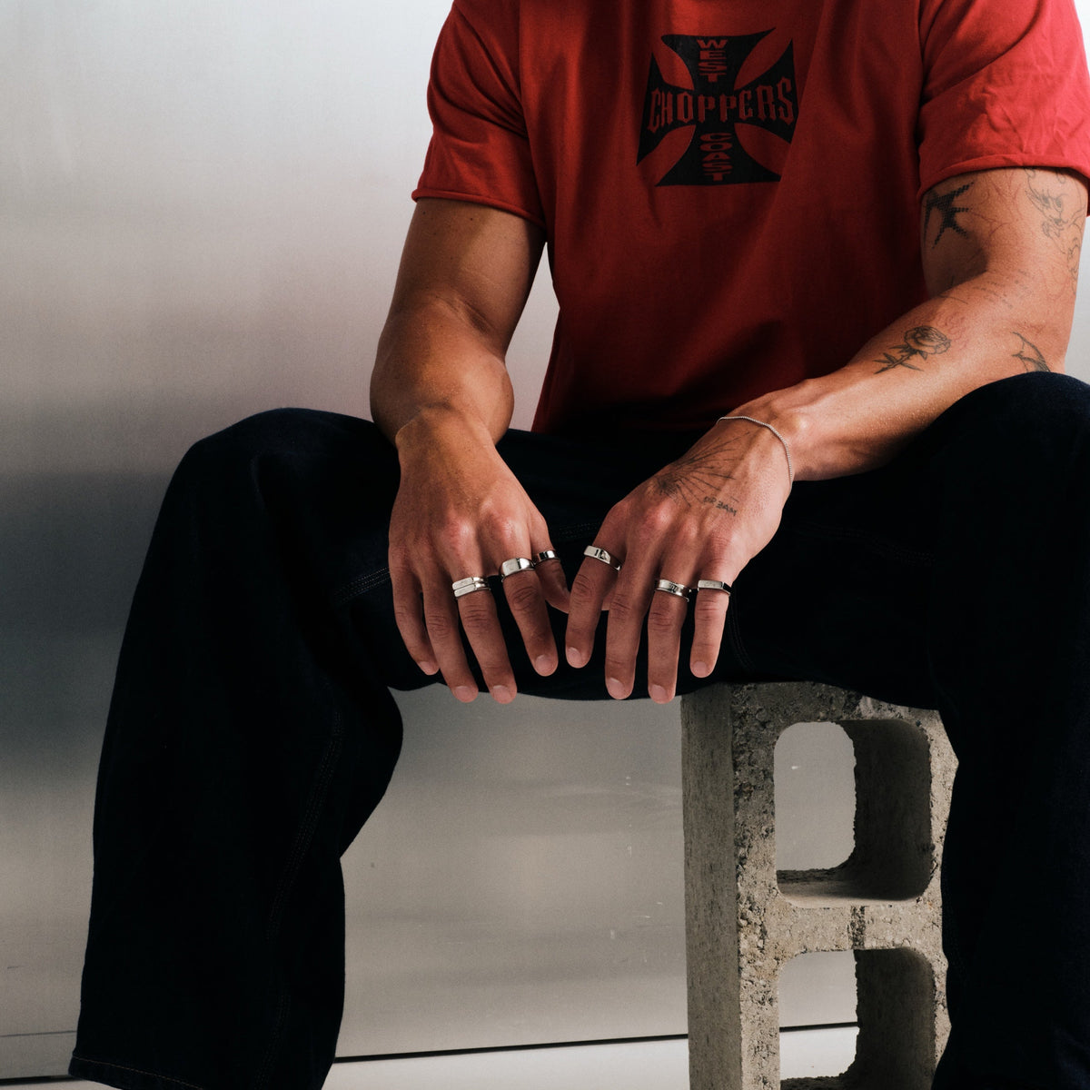 Man wearing silver finish stainless rings, seated on a concrete block, hands on his knees, with a fine steel chain bracelet