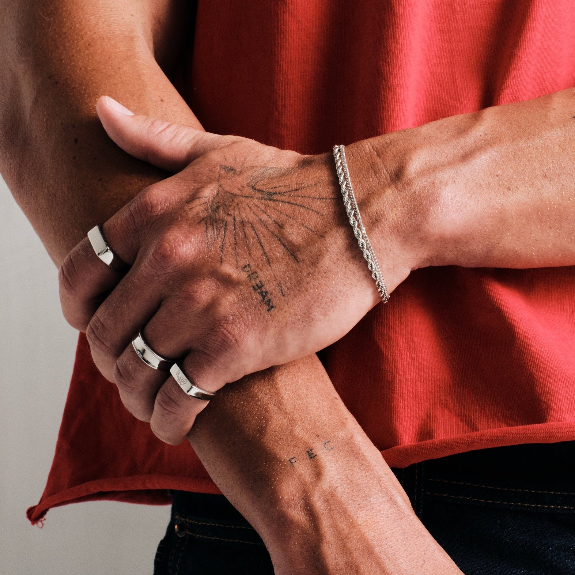 Man wearing silver finish stainless rings, hand gripping his other forearm, with a steel rope bracelet, red tee, dark jeans