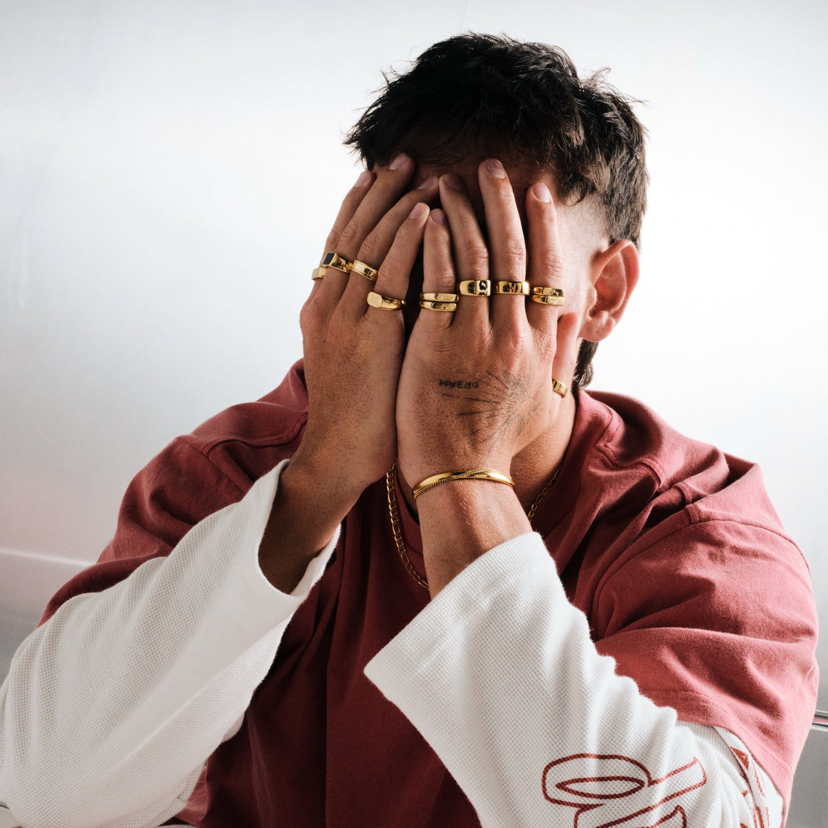 Man wearing gold stainless rings, hands over his face, with a gold chain necklace, thin gold bangle on left wrist