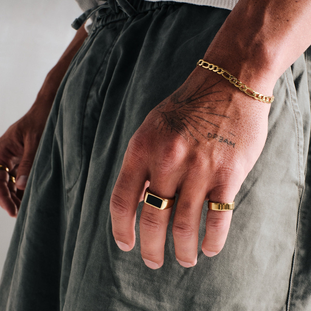 Man wearing gold stainless rings, hand by his side, with a gold Figaro bracelet on left wrist, olive pants
