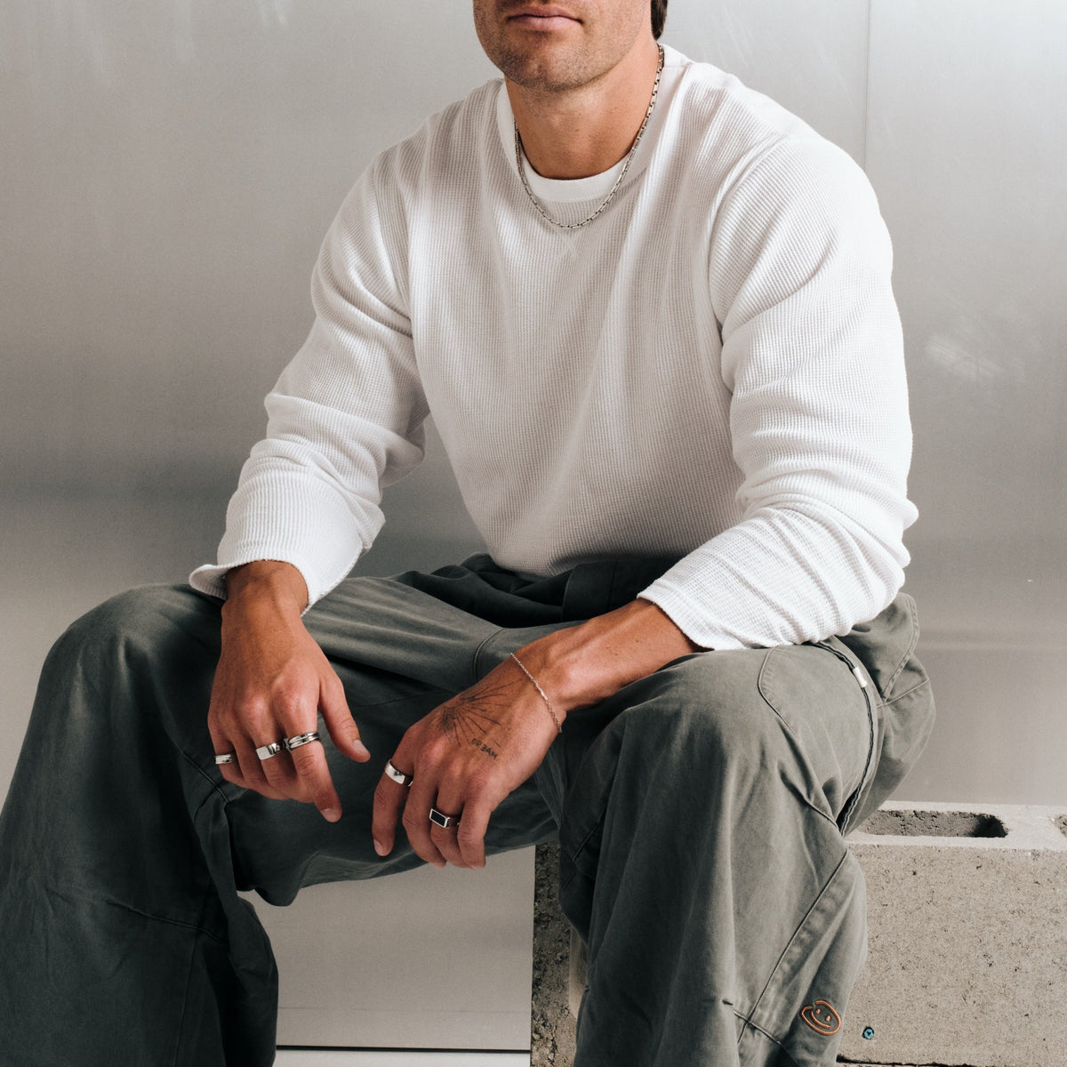 Man wearing silver finish stainless rings, seated on a concrete block, with a steel chain necklace, slim chain bracelet