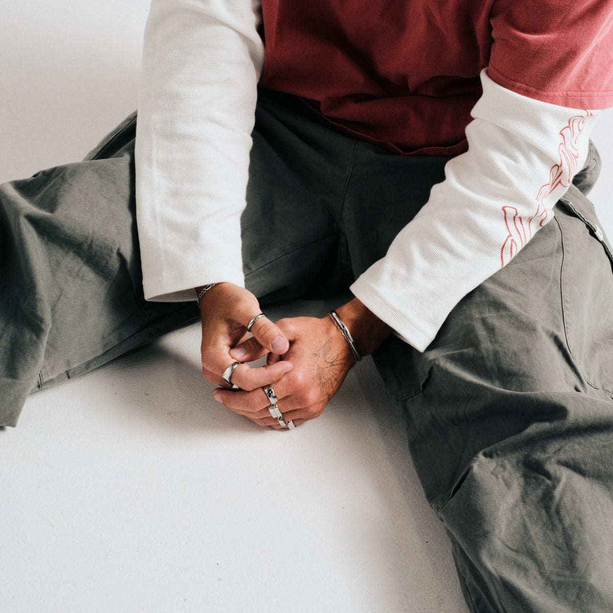 Man wearing silver finish stainless rings, seated on the floor, hands clasped, with a bangle cuff, chain bracelet
