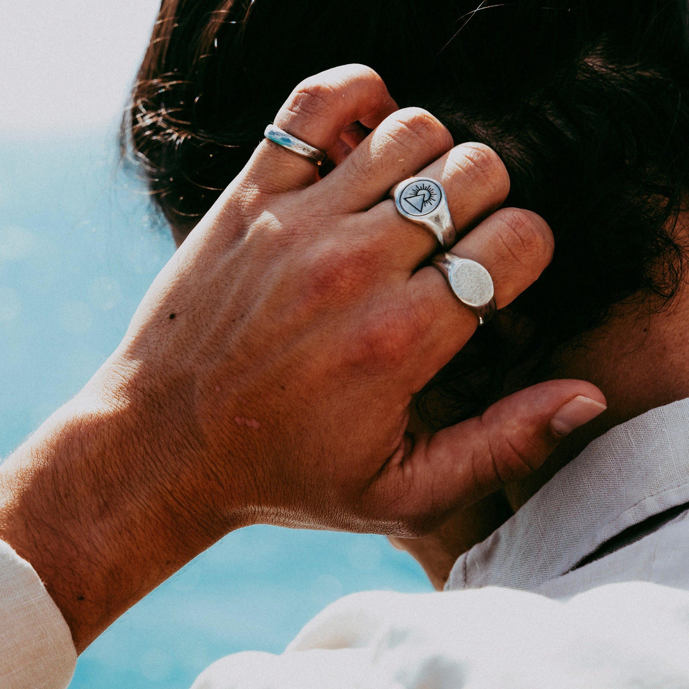 A man's hand resting on the back of his neck, showcasing the Billie Jo Signet Ring and another silver ring, with a bright, beachy background.
