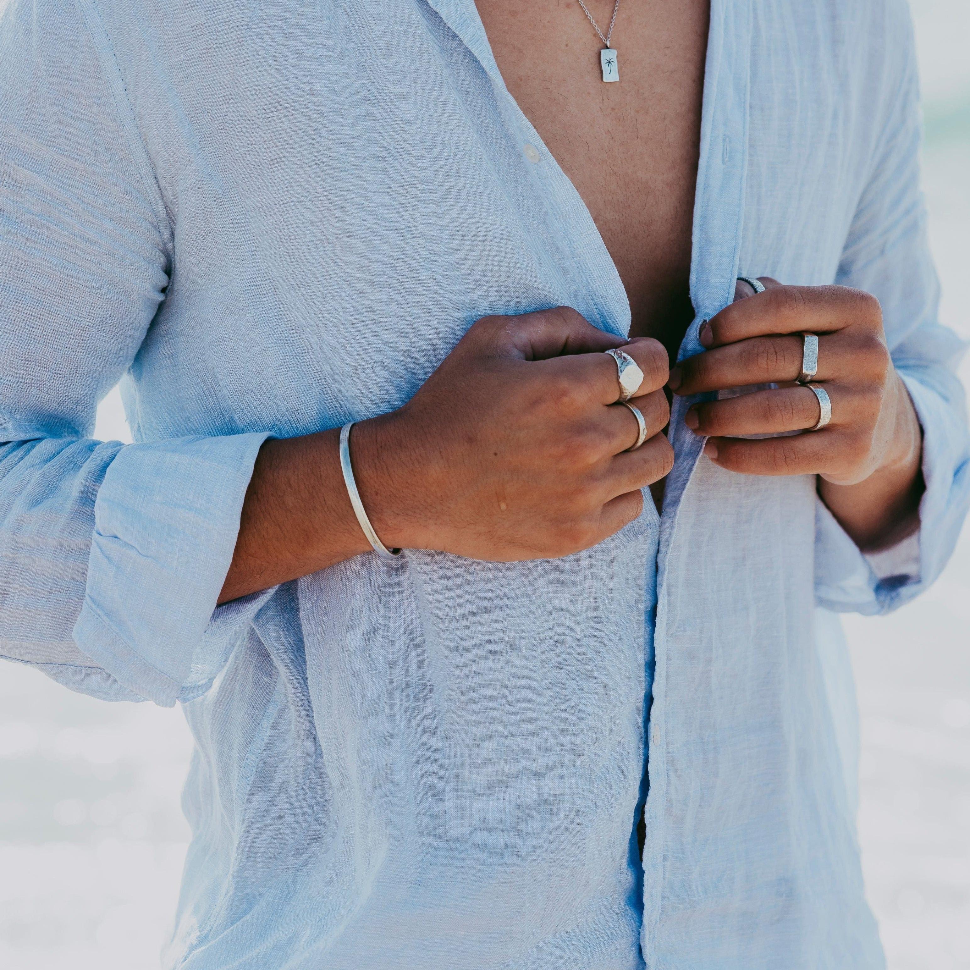 A man wearing a light blue linen shirt, unbuttoned to show a silver necklace. He is wearing the Elation Ring and other silver jewellery from Billie Jo.