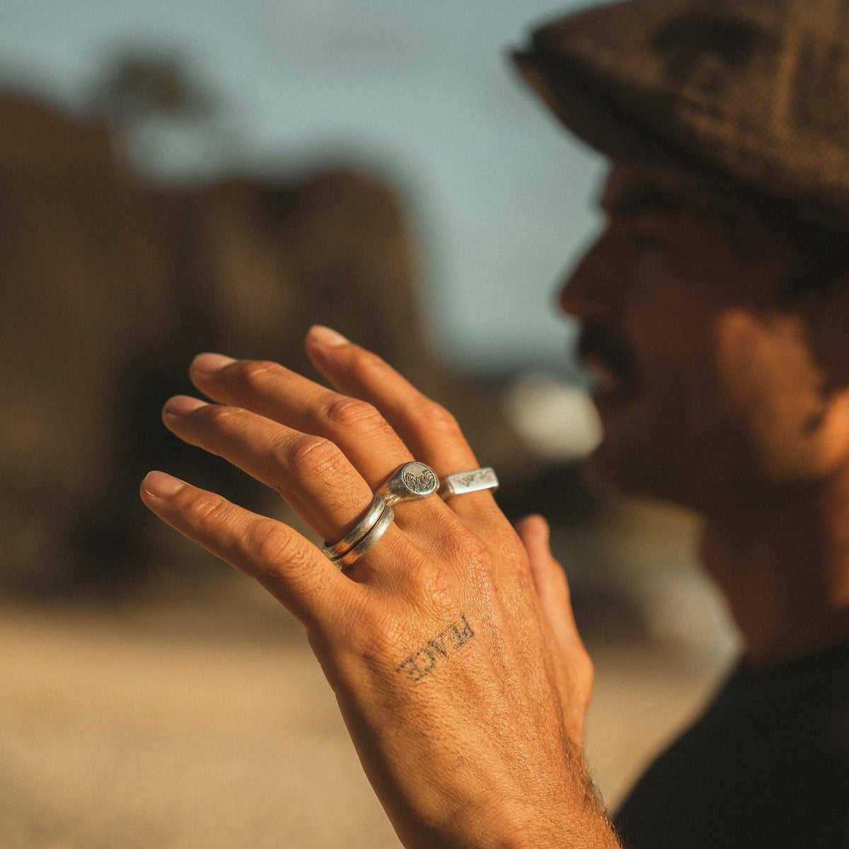 A man's hand with a 'PEACE' tattoo, wearing the Billie Jo Current Ring and another silver ring, with a soft, out-of-focus background.