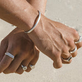 A man's hands clasped, wearing the sterling silver Elation Ring on his index finger. The shot is taken on a sandy beach, with a focus on the ring.
